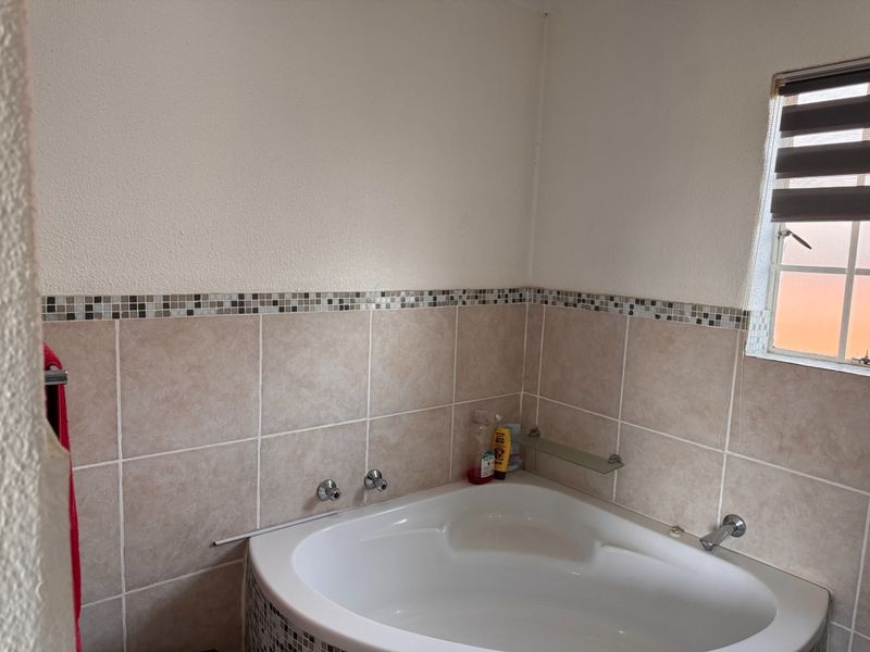 A bathroom featuring a corner bathtub with neutral wall tiling, a decorative mosaic border, and a window with a roller blind.