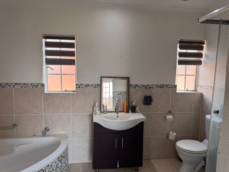 A functional bathroom featuring a corner bathtub, vanity cabinet with basin, toilet, and shower enclosure, finished with neutral wall tiles and a mosaic border.