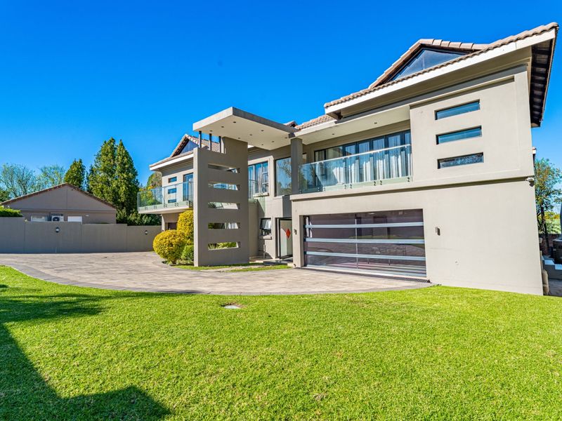 Modern double-story residence featuring a paved driveway, integrated garage, and upper-level balcony with glass balustrades, set against a well-maintained lawn.