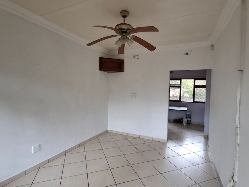 An empty living area featuring tiled flooring, a ceiling fan, and decorative cornicing, leading through to an adjacent room with diamond-pattern windows.