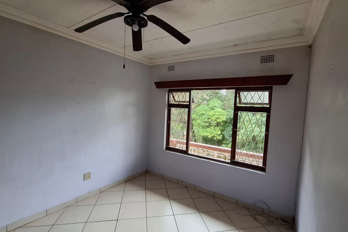An empty bedroom featuring tiled flooring, a ceiling fan, and large windows with security bars, offering a view of the surrounding greenery.