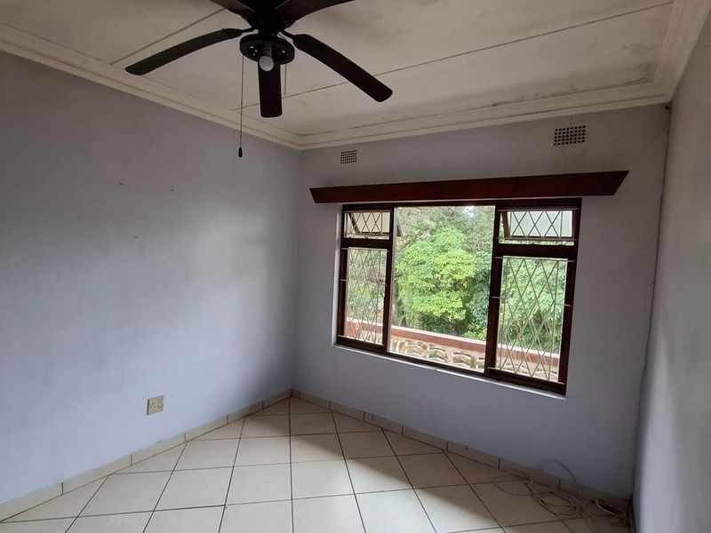 An empty bedroom featuring tiled flooring, a ceiling fan, and large windows with security bars, offering a view of the surrounding greenery.