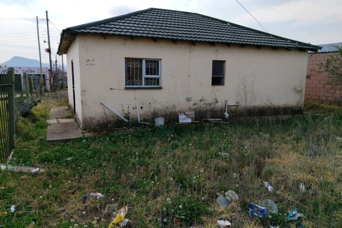 Rear view of a detached house showing significant exterior wear, including damaged wall finishes, exposed plumbing, and a neglected garden area.