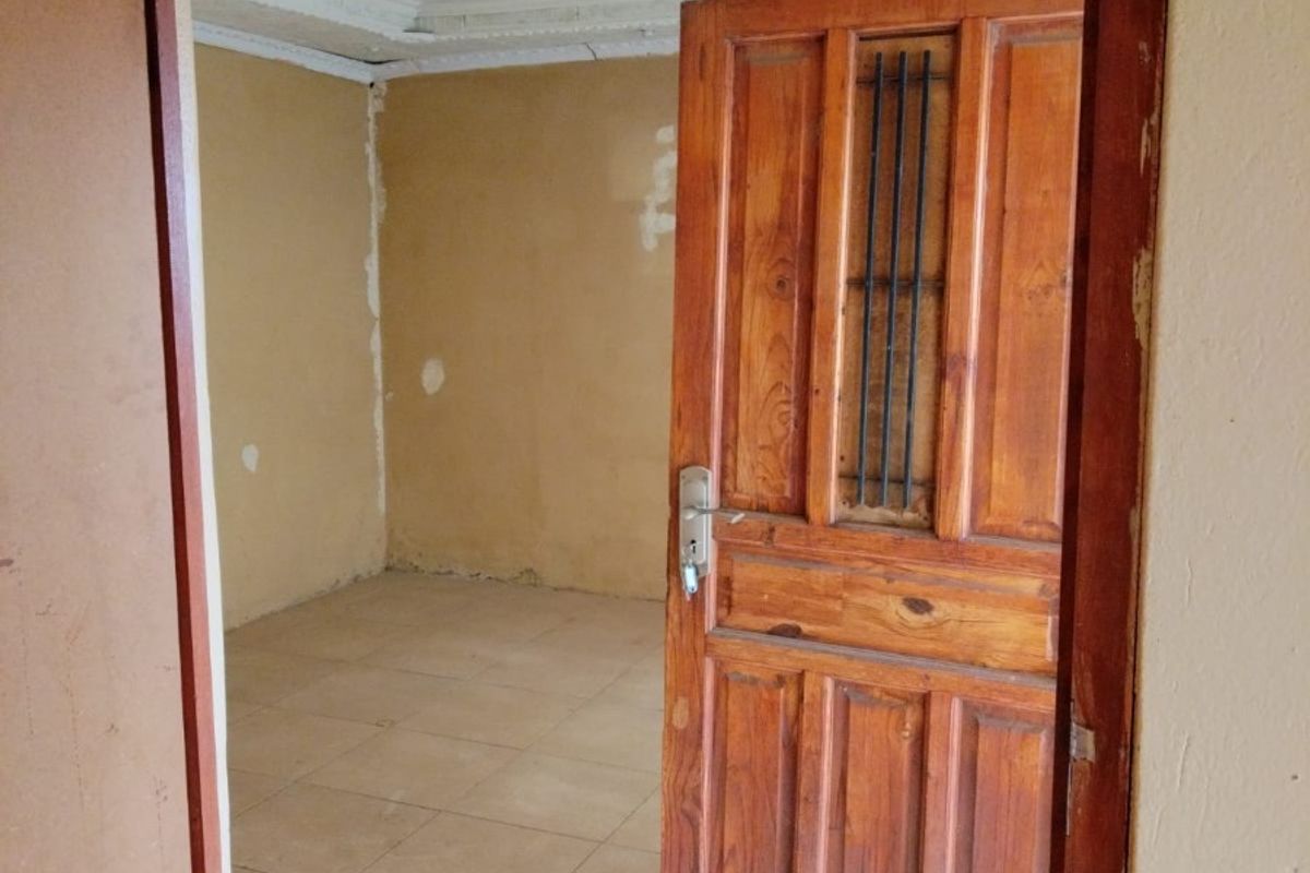 An empty bedroom featuring tiled flooring, a coved ceiling, and a wooden door with an integrated security grille, requiring cosmetic updates and wall repairs.