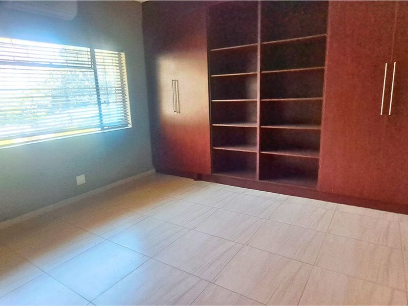 A spacious bedroom featuring tiled flooring, built-in wooden cupboards with open shelving, and venetian blinds.
