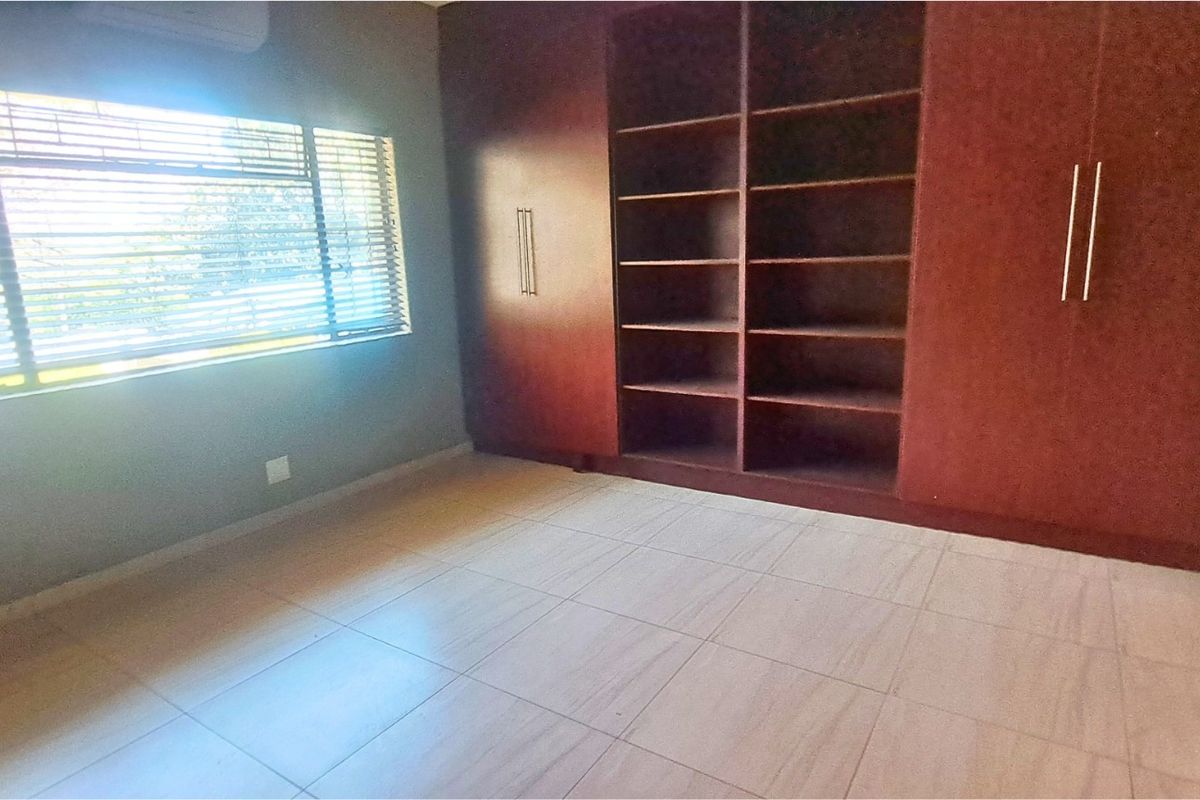 A spacious bedroom featuring tiled flooring, built-in wooden cupboards with open shelving, and venetian blinds.