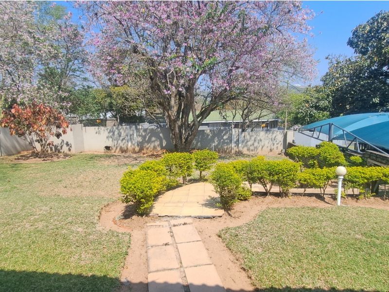 A spacious garden featuring a paved walkway, established trees, perimeter walling, and a shaded outdoor area.
