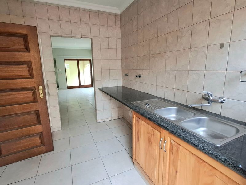 A functional scullery featuring a double stainless steel sink, granite countertops, wooden cabinetry, and neutral ceramic tiling throughout.