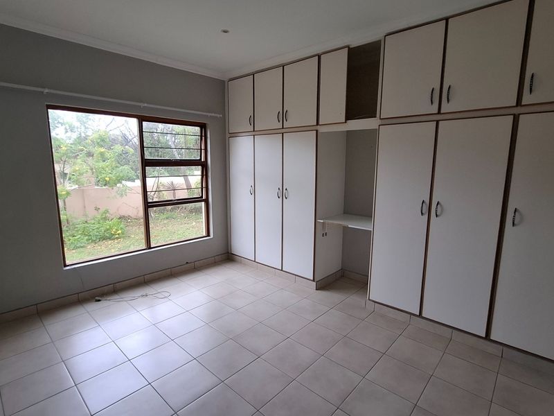 A spacious, empty bedroom featuring neutral tiled flooring, a large window providing natural light, and extensive built-in cabinetry with an integrated desk area.