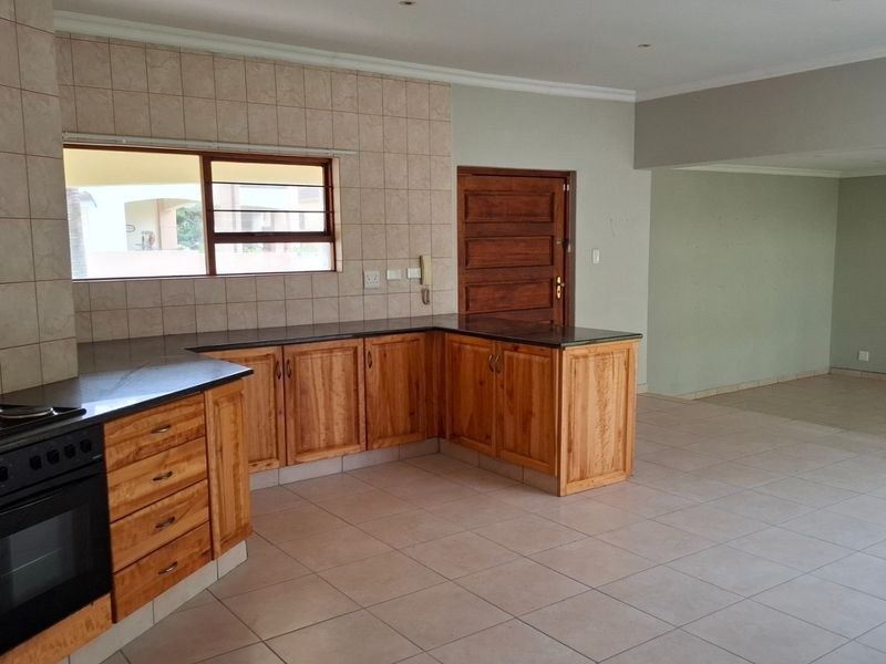 A spacious kitchen featuring wooden cabinetry, granite countertops, and a built-in oven, seamlessly flowing into an open-plan living area with neutral tiled flooring.
