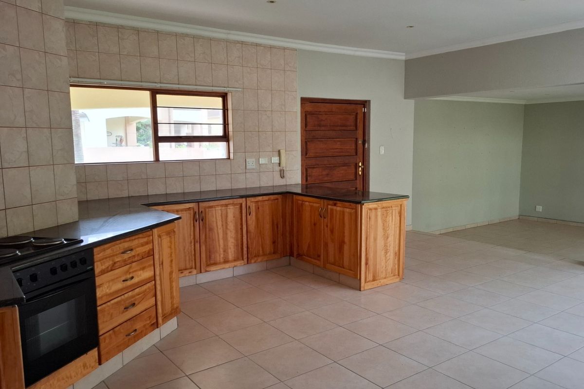 A spacious kitchen featuring wooden cabinetry, granite countertops, and a built-in oven, seamlessly flowing into an open-plan living area with neutral tiled flooring.