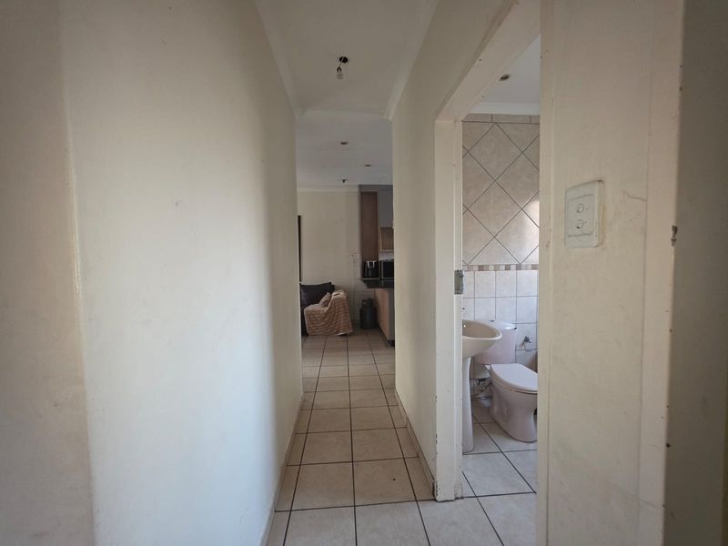 A narrow hallway with tiled flooring leading to an adjacent bathroom and open-plan living area, showing signs of wear and requiring cosmetic updates.