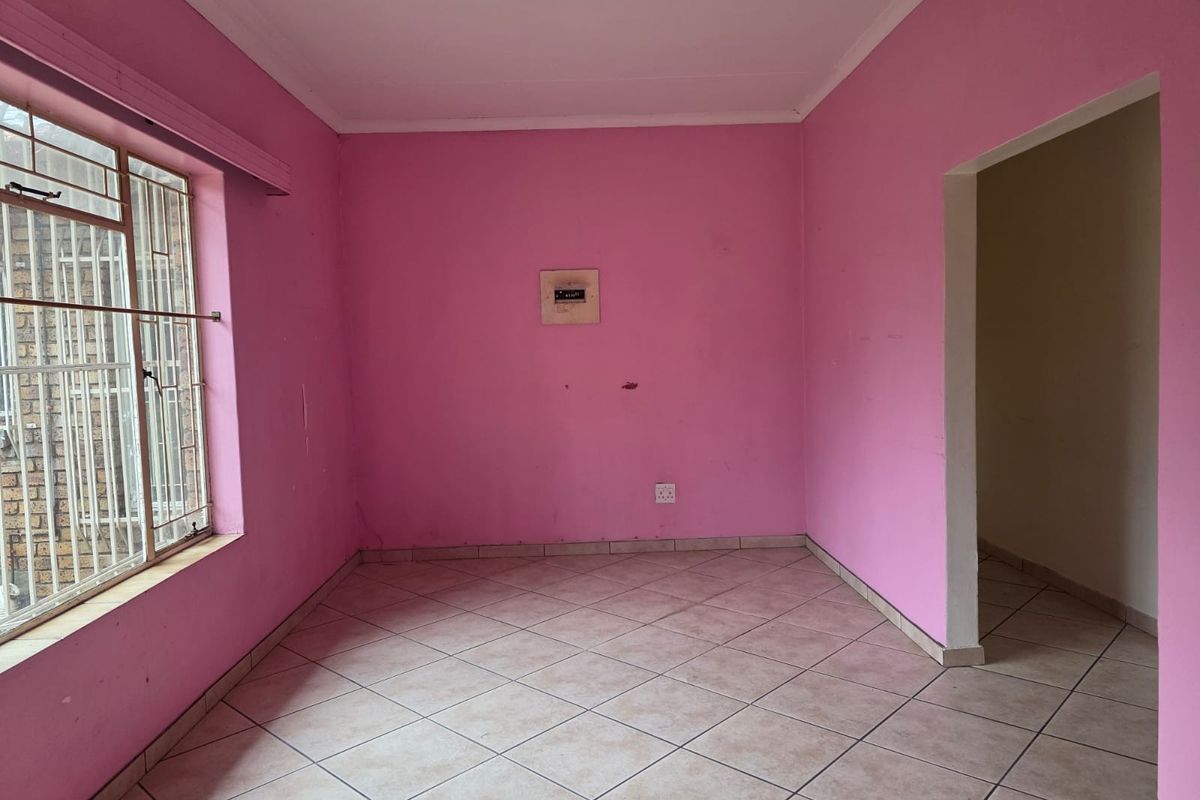 An empty living room featuring tiled flooring, security bars on the window, and an electrical distribution board, requiring cosmetic updates to the wall finishes.