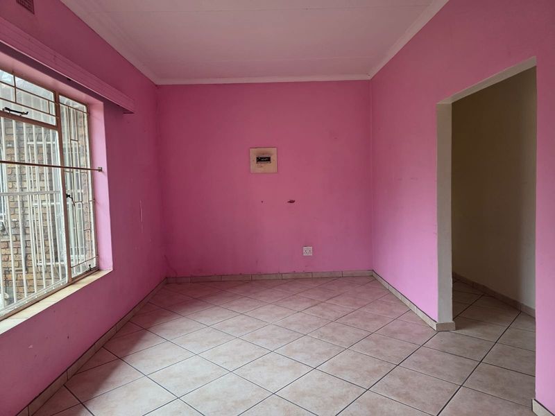 An empty living room featuring tiled flooring, security bars on the window, and an electrical distribution board, requiring cosmetic updates to the wall finishes.