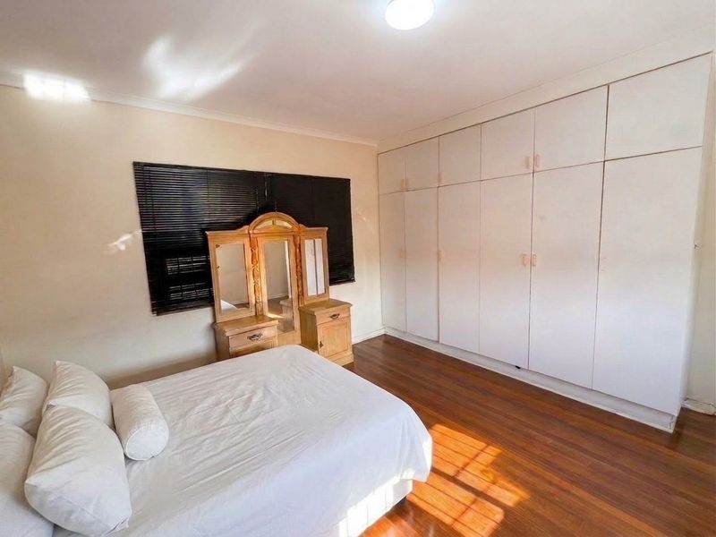 A bedroom featuring laminate flooring, extensive floor-to-ceiling built-in cupboards, and dark venetian blinds, offering a functional and well-lit living space.