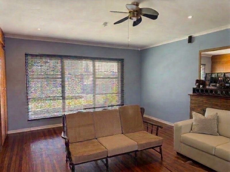 A living room featuring hardwood flooring, a brick fireplace, and a ceiling fan, offering a comfortable space with natural light from large windows.