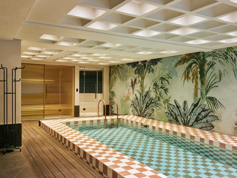 A luxurious indoor pool area featuring a decorative mosaic wall, checkered tile surround, timber decking, and a built-in sauna.