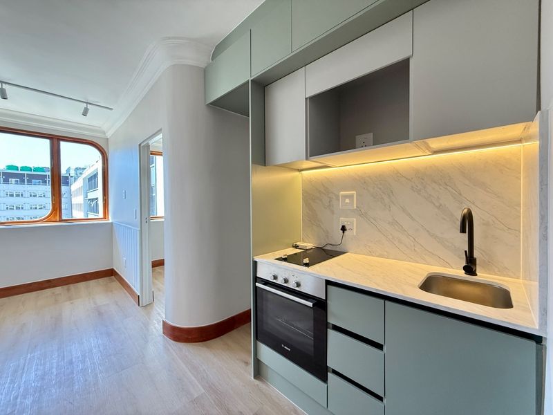 Modern, compact kitchen featuring sage green cabinetry, marble-look splashback, integrated oven and hob, and sleek matte black fixtures in a bright, open-plan apartment setting.