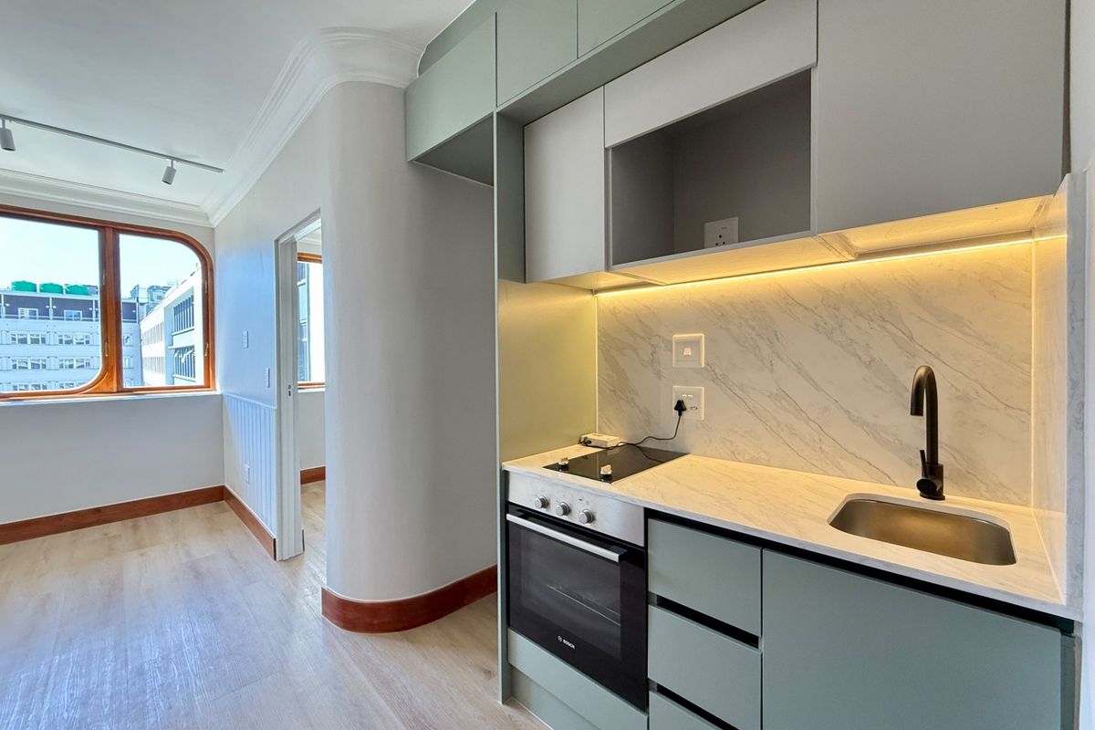 Modern, compact kitchen featuring sage green cabinetry, marble-look splashback, integrated oven and hob, and sleek matte black fixtures in a bright, open-plan apartment setting.