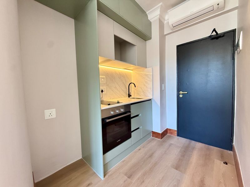 A modern, compact kitchen featuring sage green cabinetry, stone countertops, integrated oven and hob, and light wood-look flooring in a well-maintained apartment.