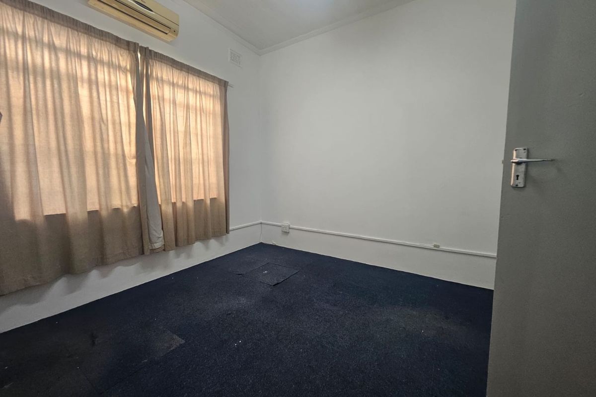 An empty office space featuring dark carpet flooring, a wall-mounted air conditioning unit, and surface-mounted electrical conduits.