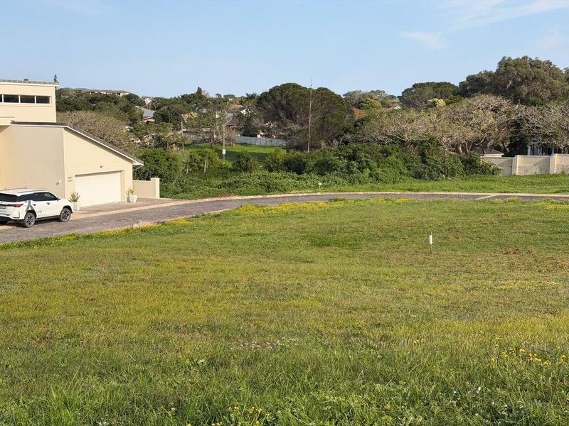 A vacant residential plot featuring a gentle slope, established grass, and a paved driveway leading to an adjacent property.