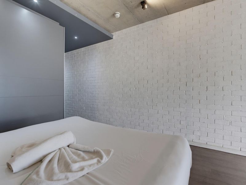 Modern bedroom featuring an exposed concrete ceiling, painted brick feature wall, and sleek built-in cabinetry with recessed lighting.