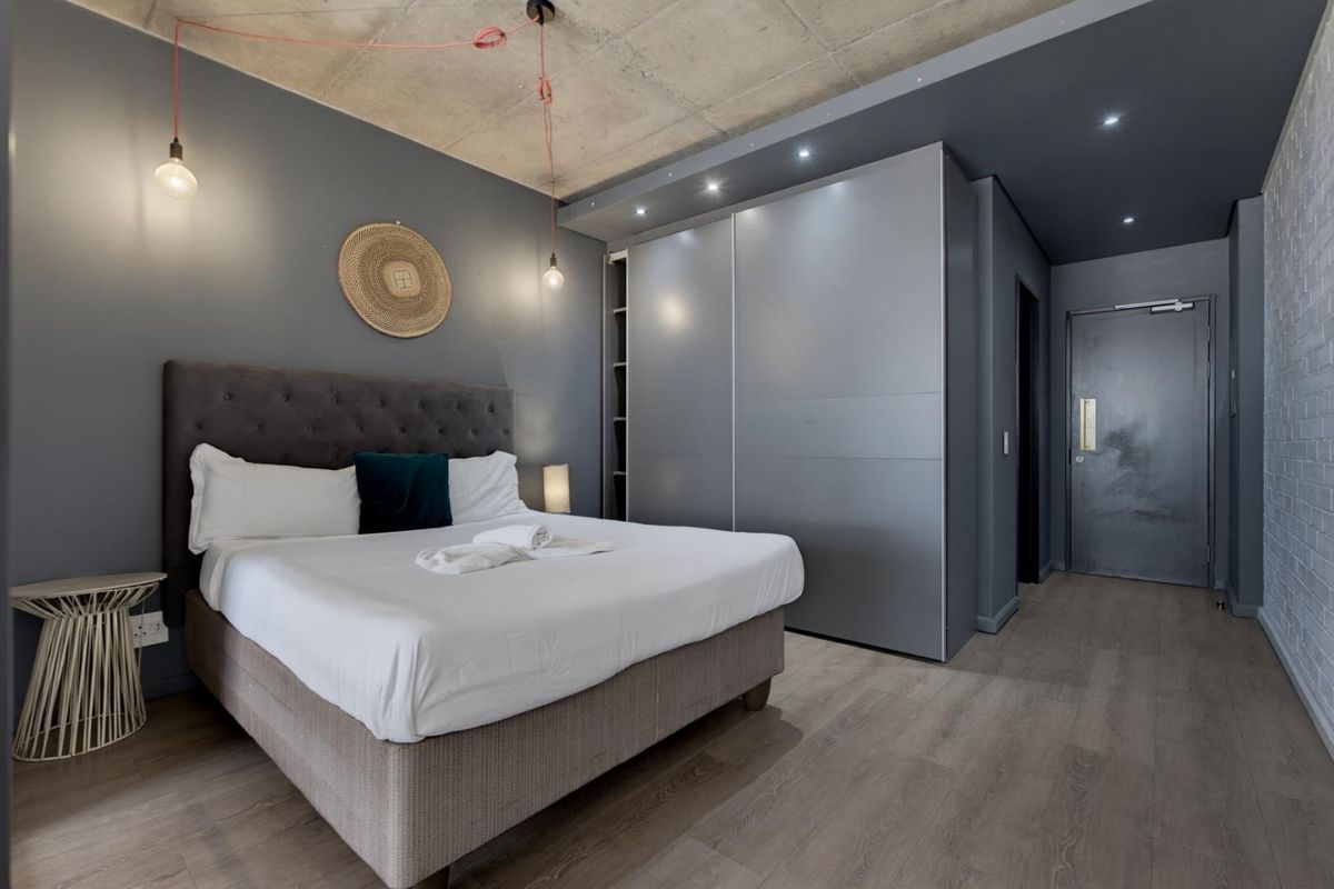 Modern bedroom featuring an exposed concrete ceiling, sleek built-in sliding wardrobes, laminate flooring, and contemporary lighting fixtures.