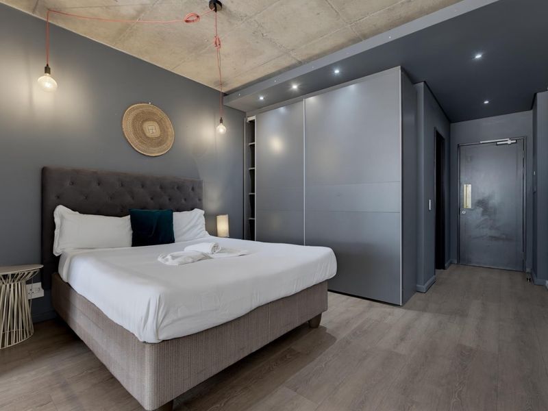 Modern bedroom featuring an exposed concrete ceiling, sleek built-in sliding wardrobes, laminate flooring, and contemporary lighting fixtures.