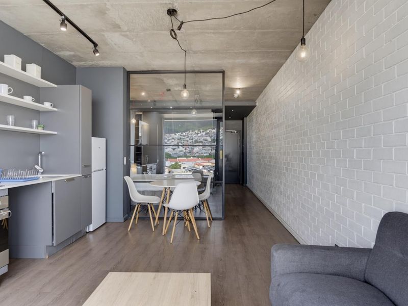 Modern open-plan living area featuring an exposed concrete ceiling, painted brick feature wall, laminate flooring, and a sleek kitchen with integrated appliances.