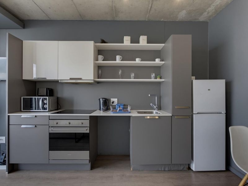 Modern kitchen featuring grey cabinetry, white upper cabinets, open shelving, integrated oven and hob, and an exposed concrete ceiling.