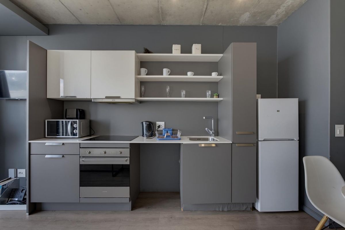 Modern kitchen featuring grey cabinetry, white upper cabinets, open shelving, integrated oven and hob, and an exposed concrete ceiling.