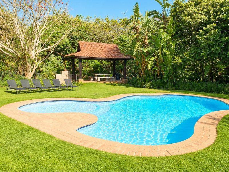 A well-maintained swimming pool area featuring a curved brick coping, lush garden surroundings, and a covered gazebo with a built-in braai.