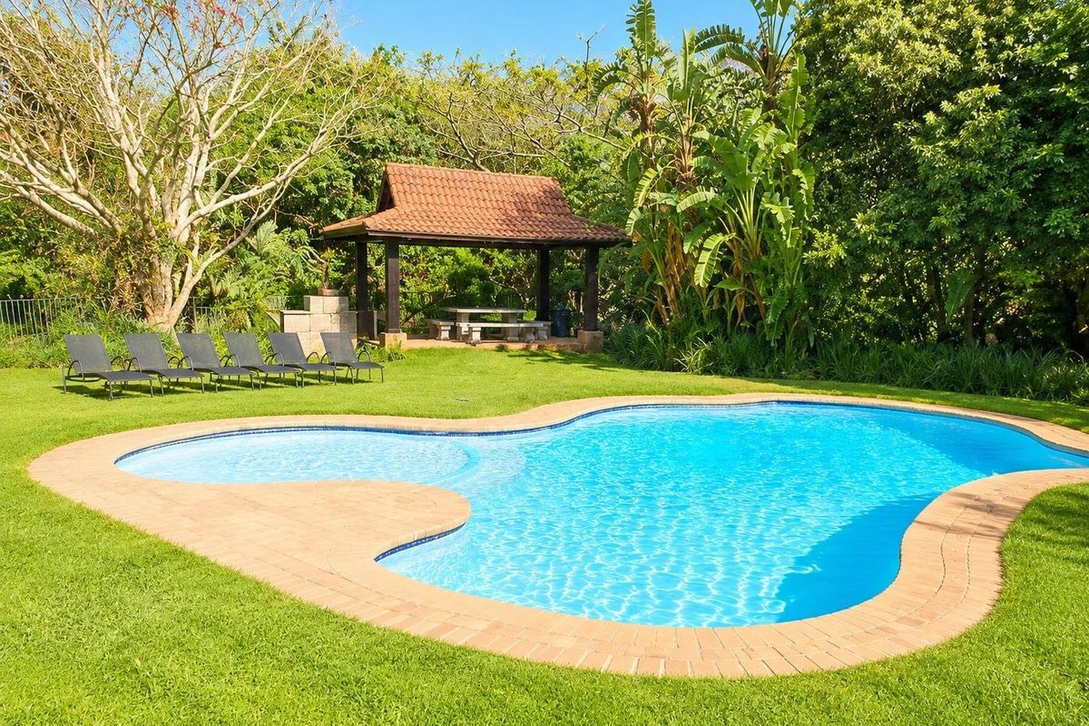 A well-maintained swimming pool area featuring a curved brick coping, lush garden surroundings, and a covered gazebo with a built-in braai.