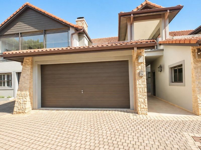 Front elevation of a modern house featuring a double garage, stone-clad pillars, tiled roof, and a paved driveway leading to the entrance.