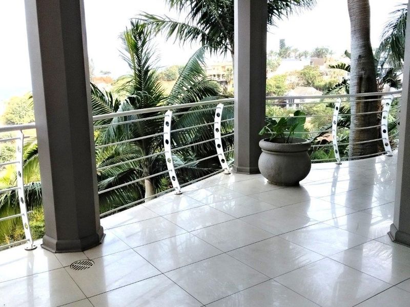 A spacious covered balcony featuring white tiled flooring, structural columns, and a modern metal balustrade, offering an open view of the surrounding landscape.