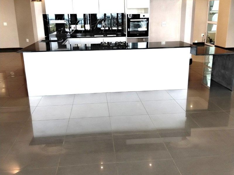 Modern open-plan kitchen featuring a large island, sleek white cabinetry, black stone countertops, built-in oven, and polished tiled flooring.
