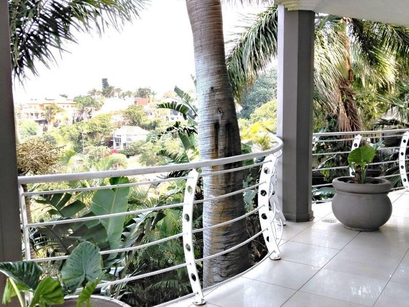 A spacious covered balcony featuring tiled flooring, a modern metal balustrade, and support columns, offering a pleasant view of the surrounding greenery.