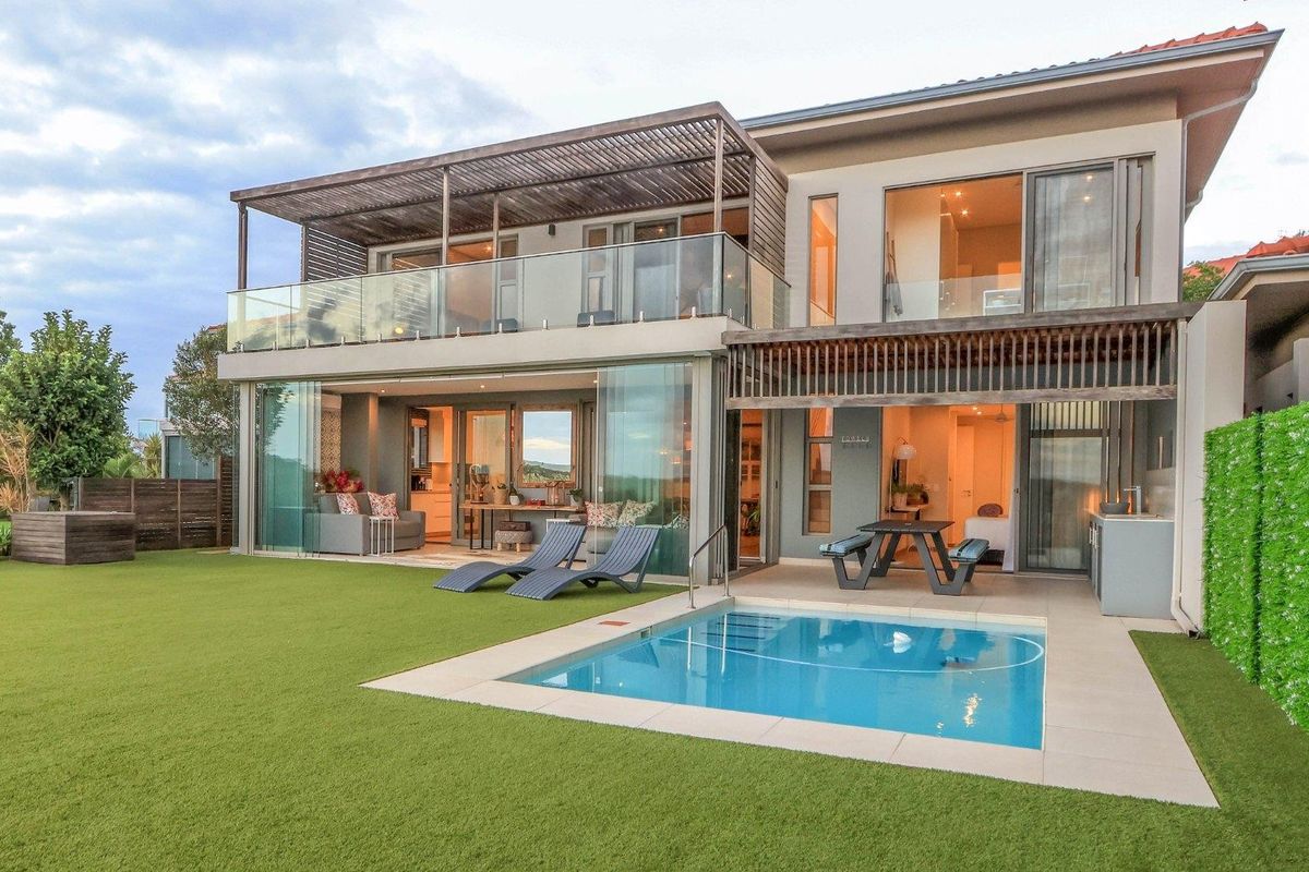 Modern double-story rear elevation featuring a swimming pool, covered patio, balcony with glass balustrades, and seamless indoor-outdoor flow through large sliding glass doors.