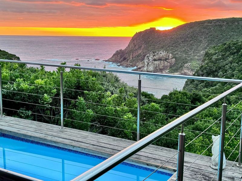 A stunning pool area featuring timber decking and modern stainless steel railing, overlooking a dramatic coastal sunset and ocean view.