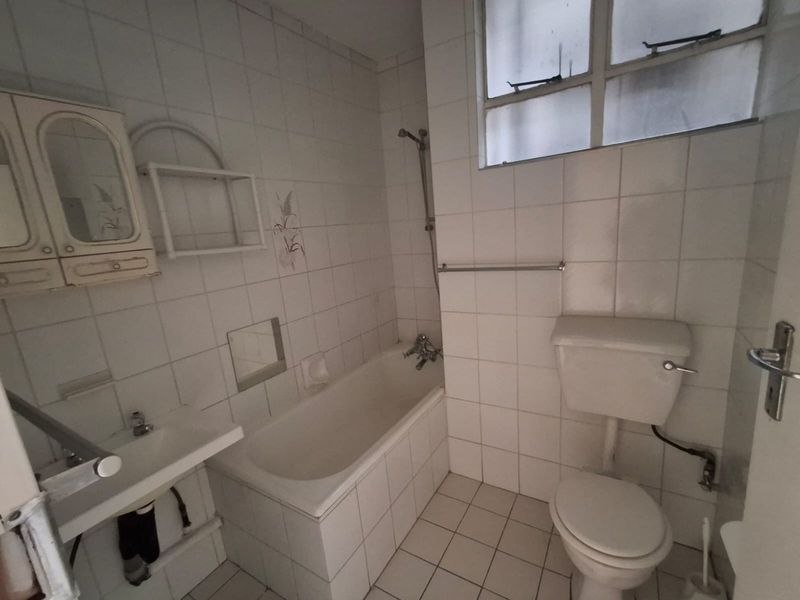 A dated bathroom featuring a standard bathtub, wall-mounted basin, and toilet with white tiled walls and flooring.