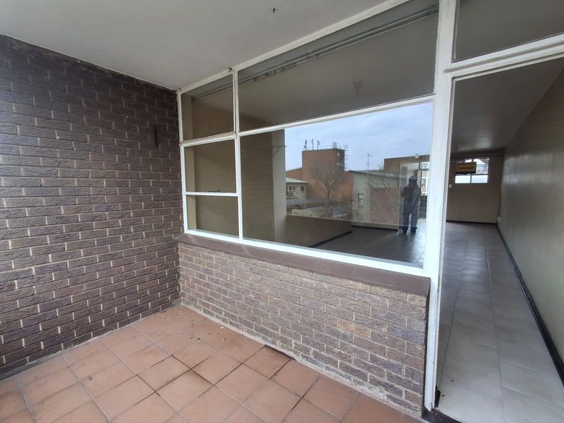 A covered balcony area featuring brick wall finishes, tiled flooring, and large glass windows overlooking the surrounding building exterior.