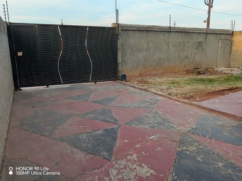 Front entrance area featuring a decorative metal sliding gate, perimeter wall with electric fencing, and a patterned paved driveway showing signs of wear.