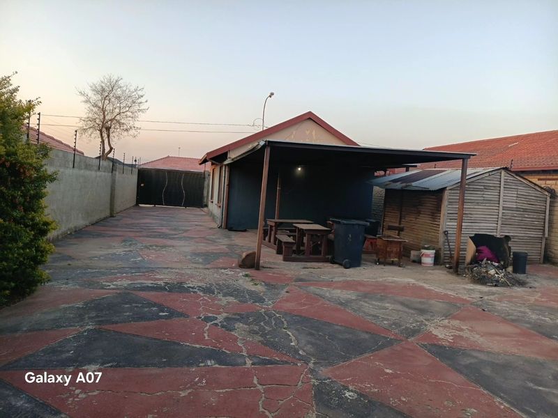 Rear exterior view featuring a large paved area, a covered patio structure, perimeter wall with electric fencing, and a timber storage shed.