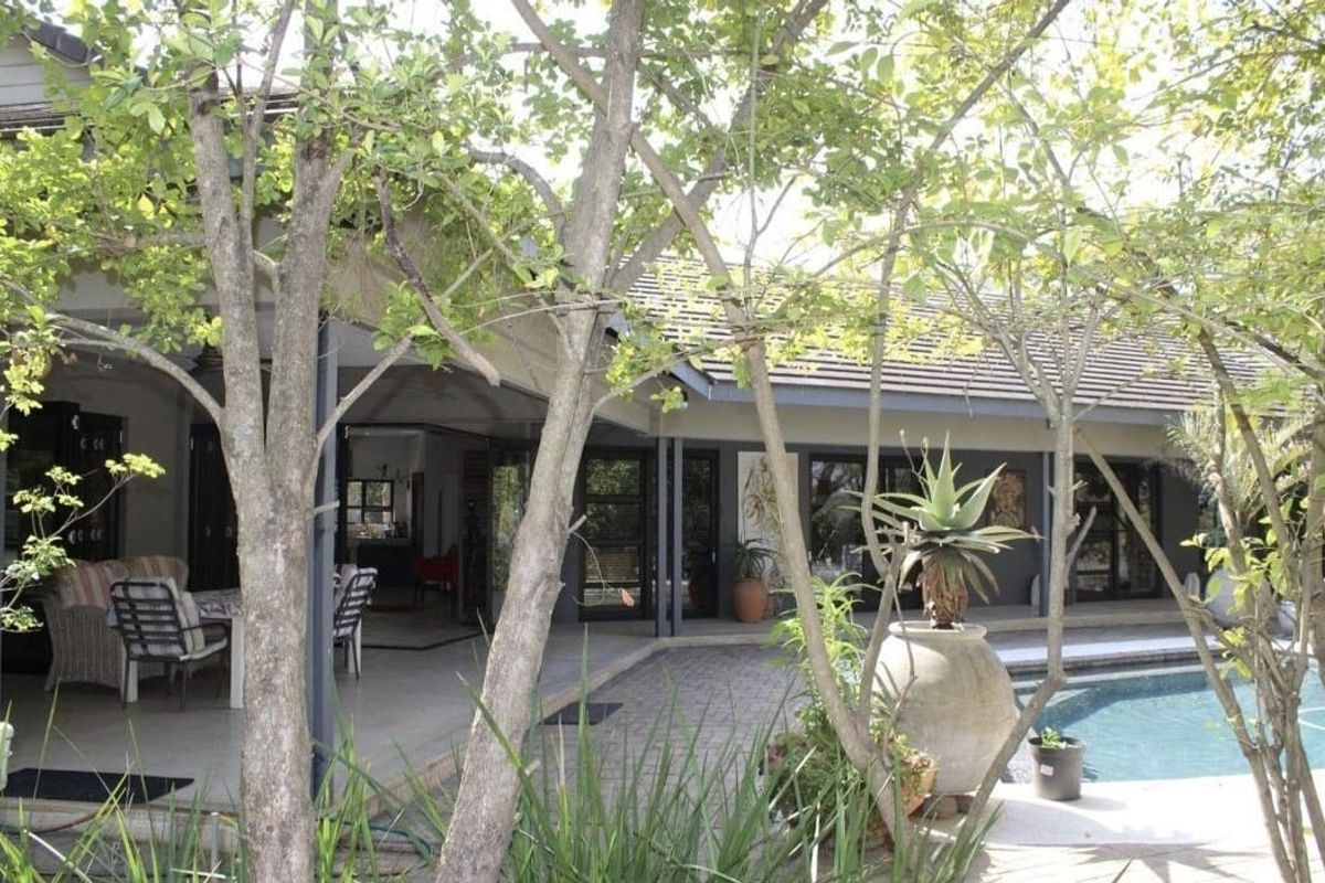A spacious rear exterior view featuring a covered patio area, paved pool deck, and a swimming pool surrounded by mature trees.