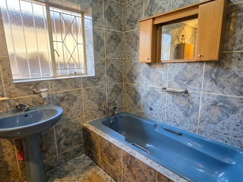A dated bathroom featuring blue sanitaryware, full-height patterned wall tiles, a pedestal basin, and a built-in bathtub with security bars on the window.