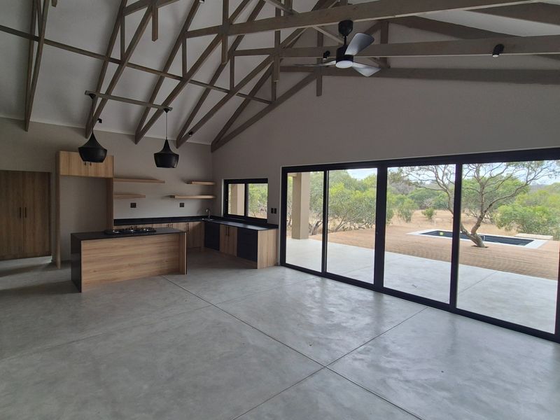 Spacious open-plan living and kitchen area featuring high vaulted ceilings with exposed timber trusses, polished concrete floors, and large sliding doors opening to the patio.