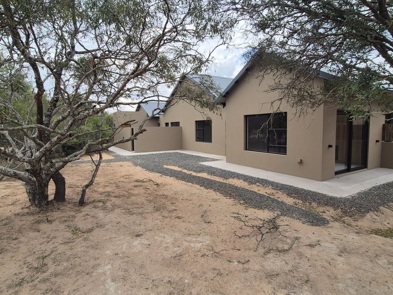 Rear view of a modern house featuring a neutral-toned facade, metal roofing, sliding glass doors, and a concrete patio area set within a natural landscape.