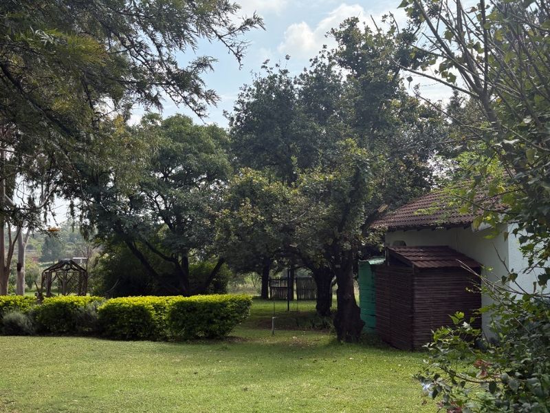 A lush, well-maintained farm garden featuring mature trees, manicured hedges, and a small outbuilding with a tiled roof.