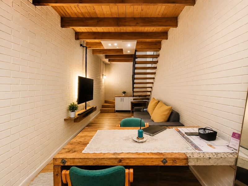 A modern open-plan living area featuring exposed timber ceiling beams, painted brick walls, and a stylish timber staircase leading to an upper level.